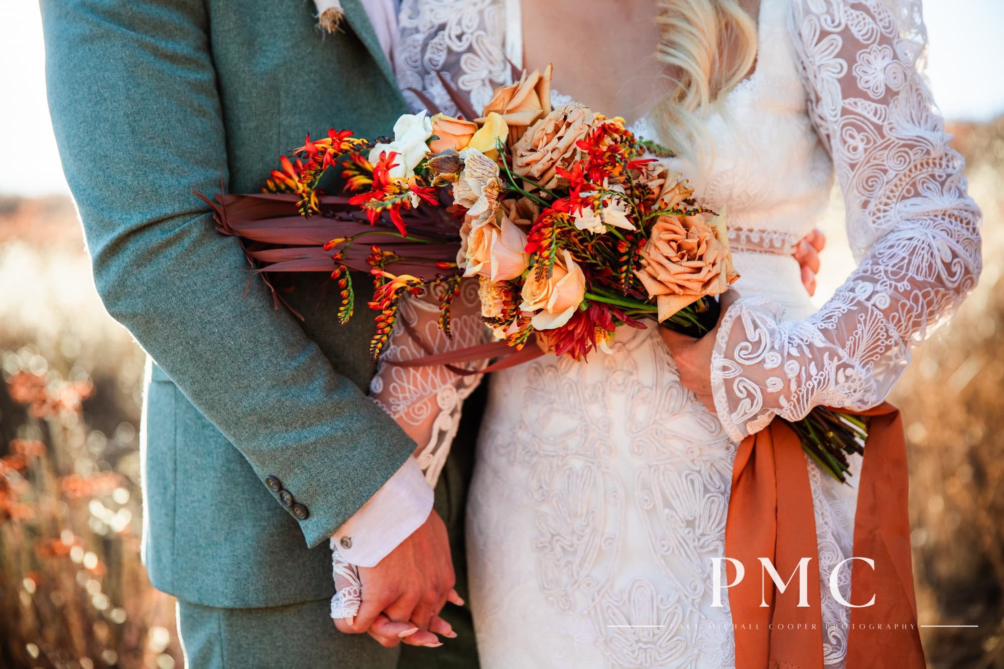 Close-up photo of a bride and groom's hands holding each other and the bride's vibrant floral bouquet.