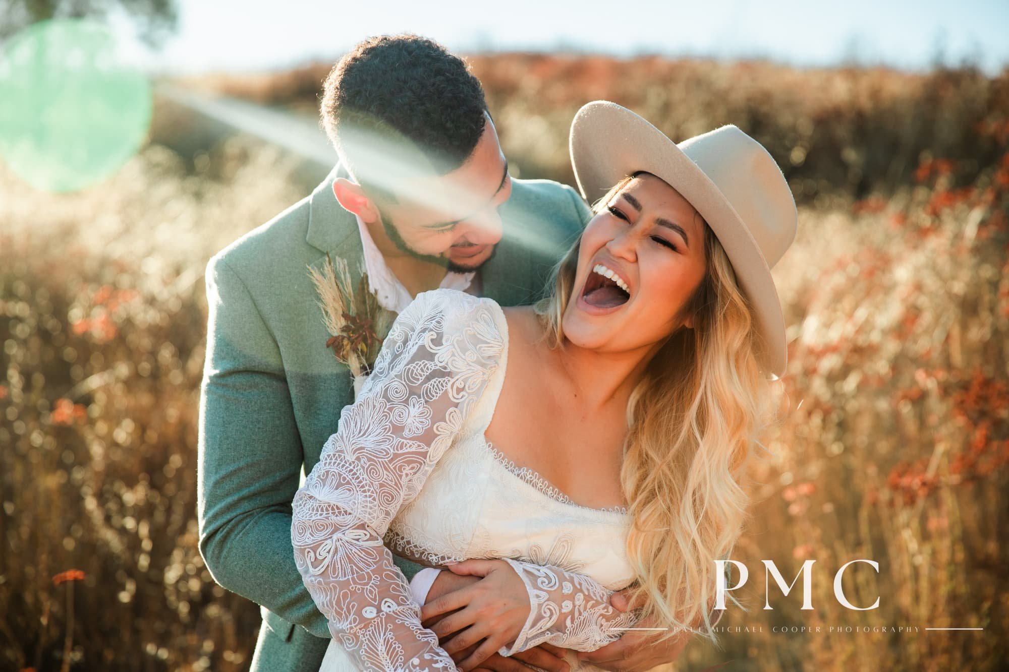 A bride in a bohemian wedding dress and a groom in a green linen suit laugh in a field in Temecula.