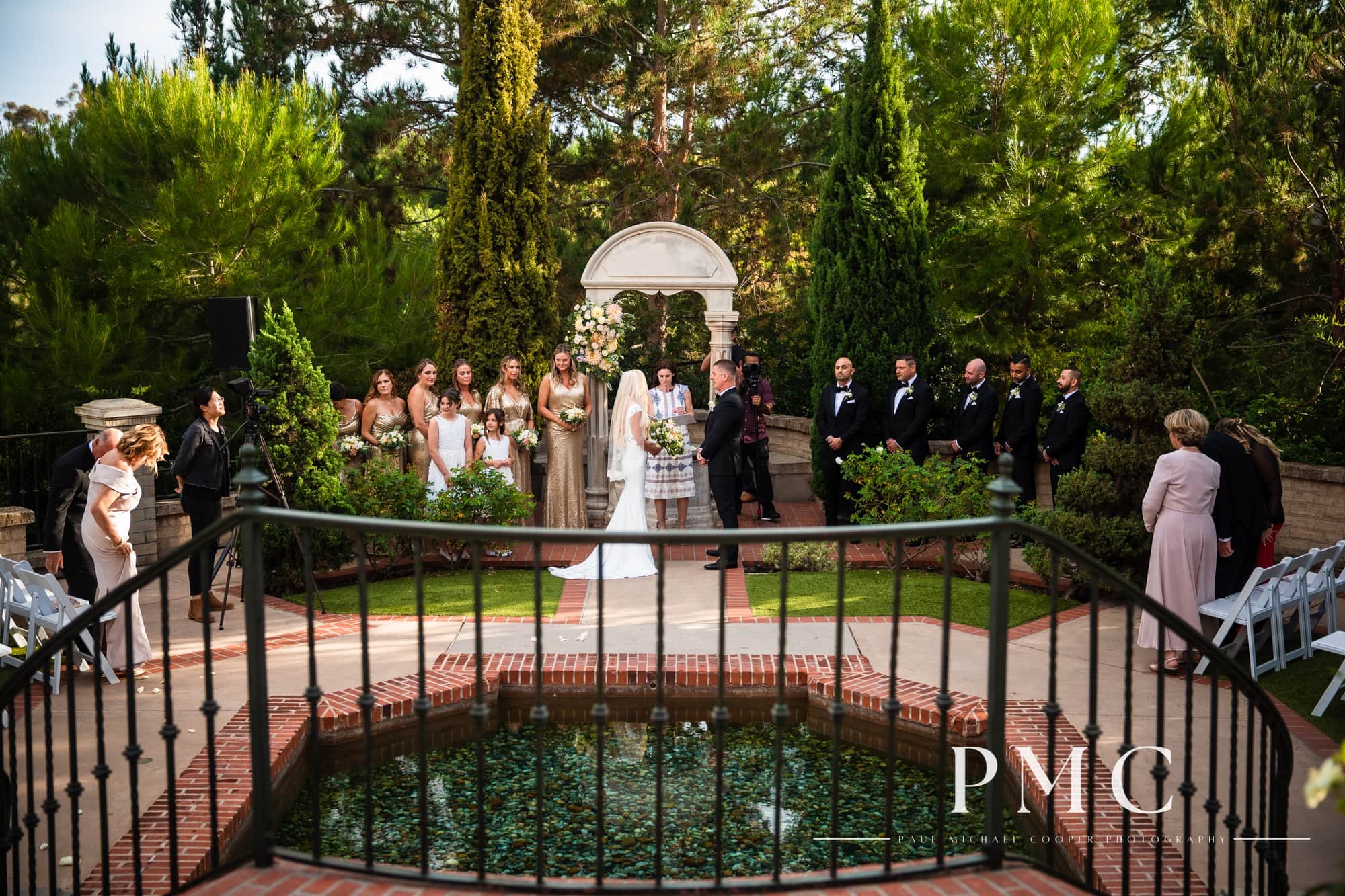A wedding ceremony in a courtyard with a fountain and surrounding green trees and foliage.