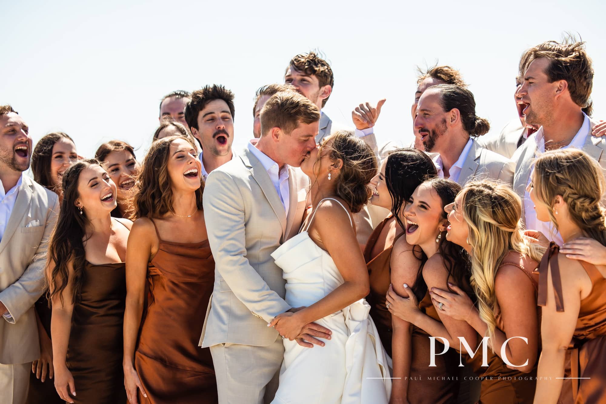 A bride and groom kiss while surrounded by their cheering wedding bridal party.