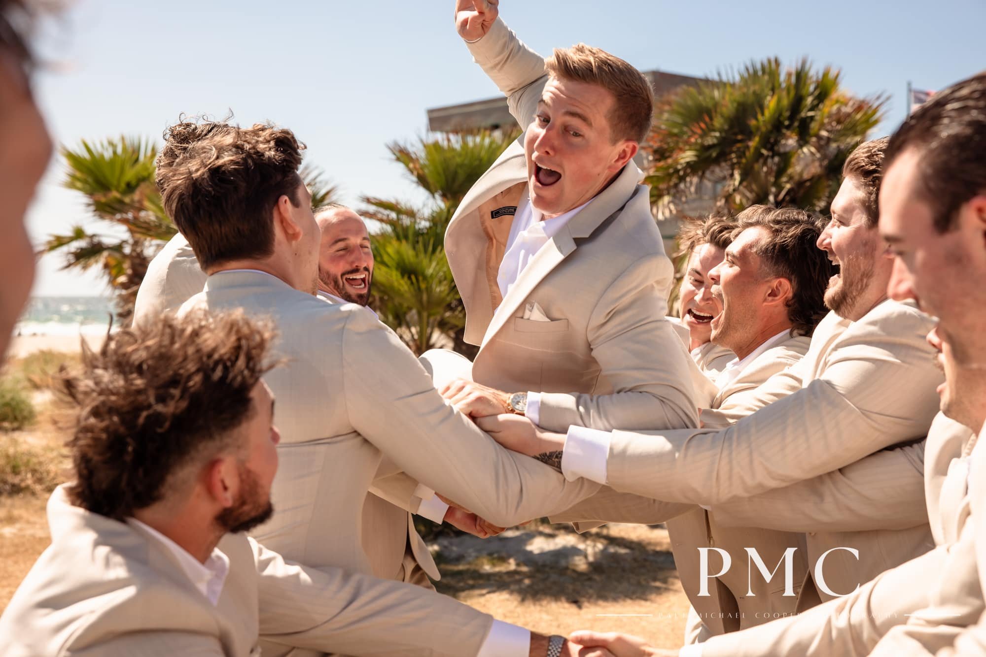 A groom leaps into the arms of his groomsmen at the beach.