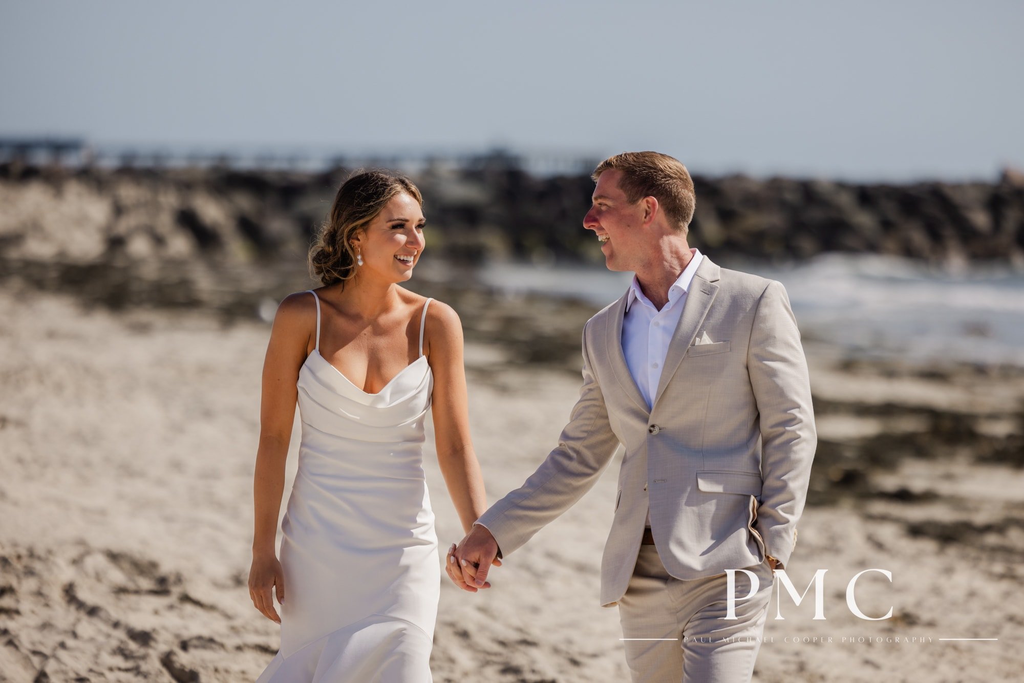 A bride and groom hold hands and smile as they walk on the beach in Mission Bay.