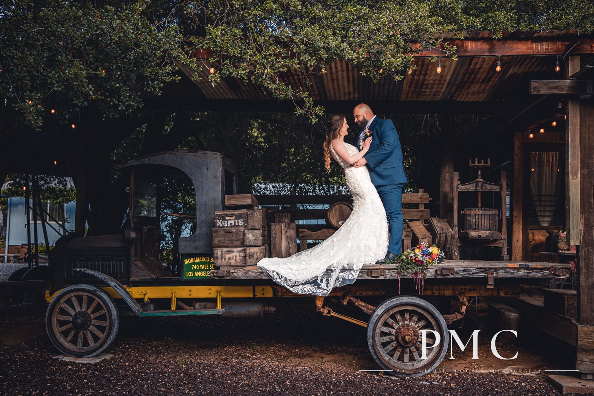 A bride and groom have a dip pose with the bride's floral bouquet on a vintage farm truck at their outdoor rustic spring wedding.