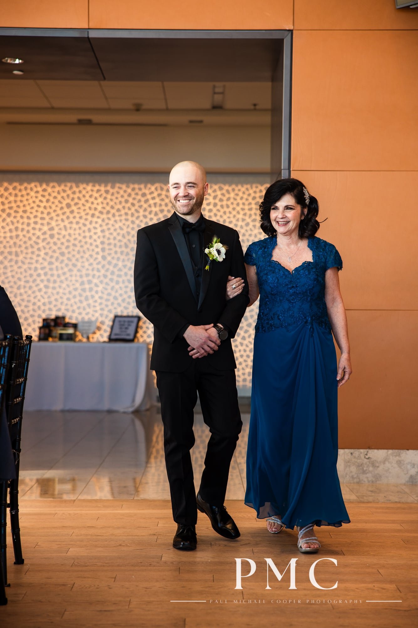 A groom smiles as he escorts his mother down the aisle during his wedding ceremony at the Ultimate Skybox in Downtown San Diego.