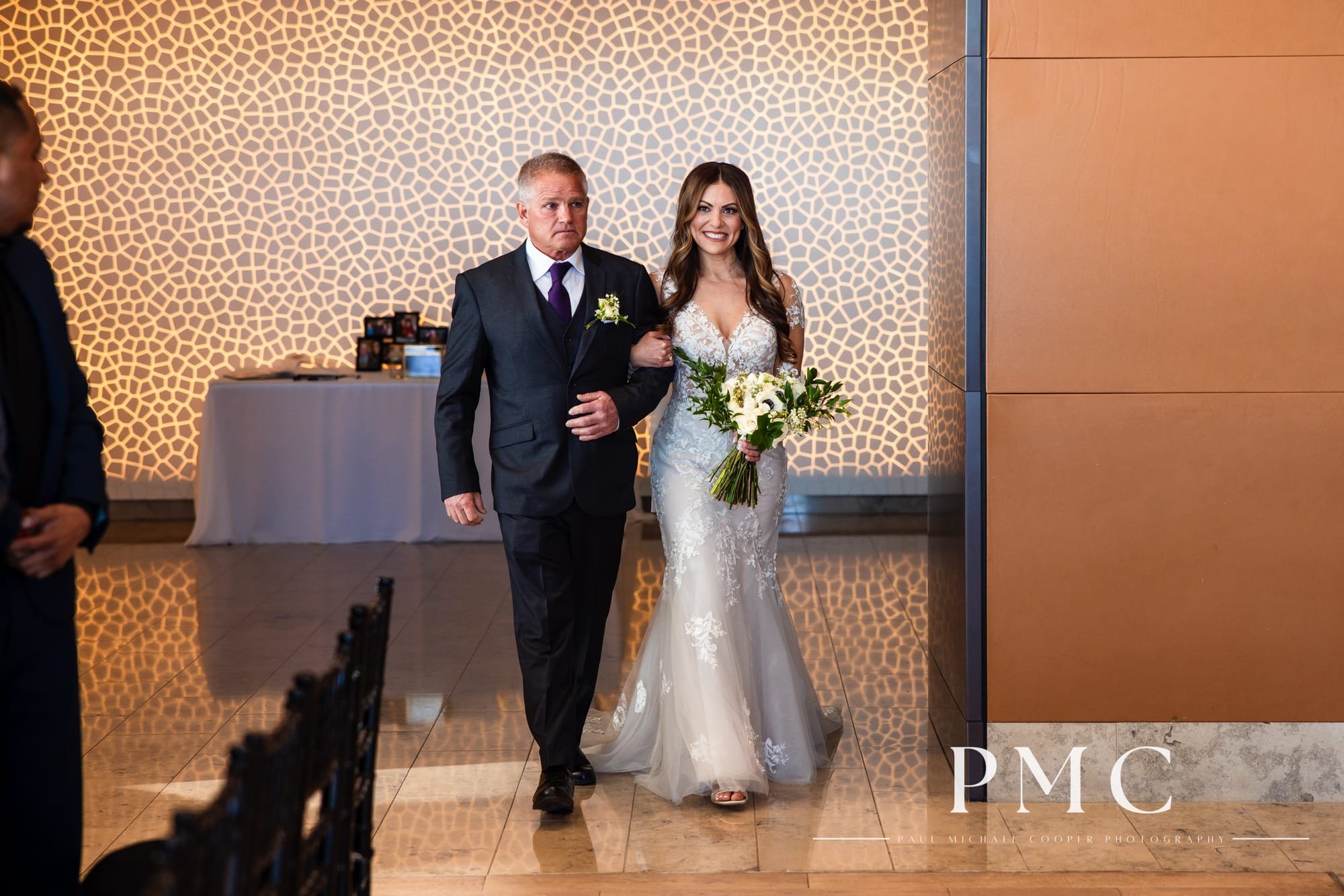 A bride smiles as she is escorted by her father down the aisle during her wedding ceremony at the Ultimate Skybox in Downtown San Diego.