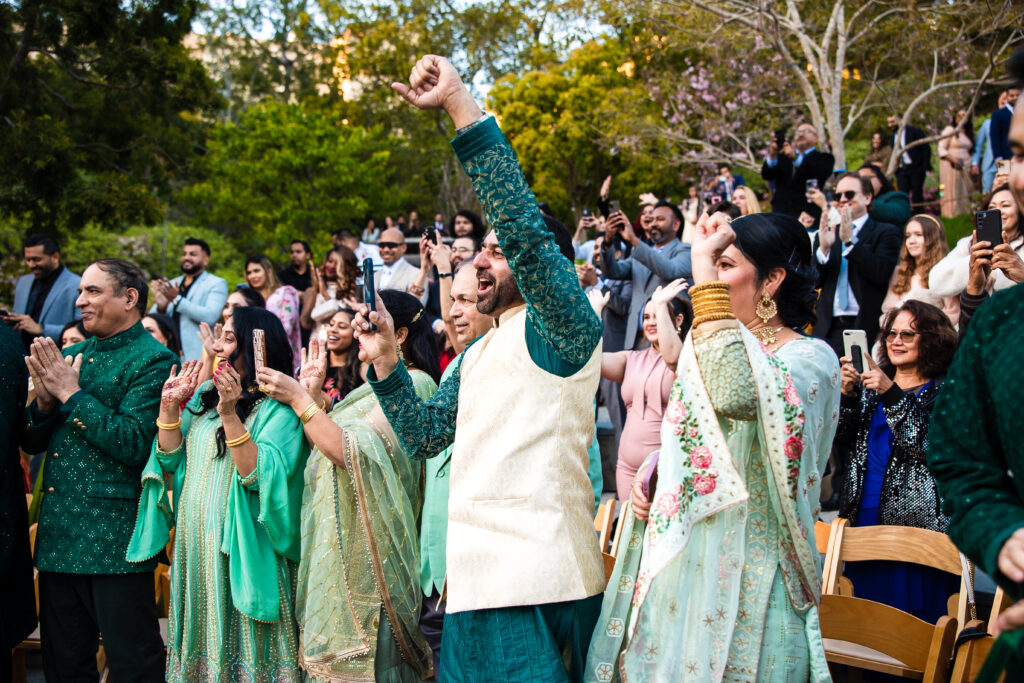 Wedding guests in traditional Indian wedding attire stand and cheer at an outdoor spring wedding ceremony.