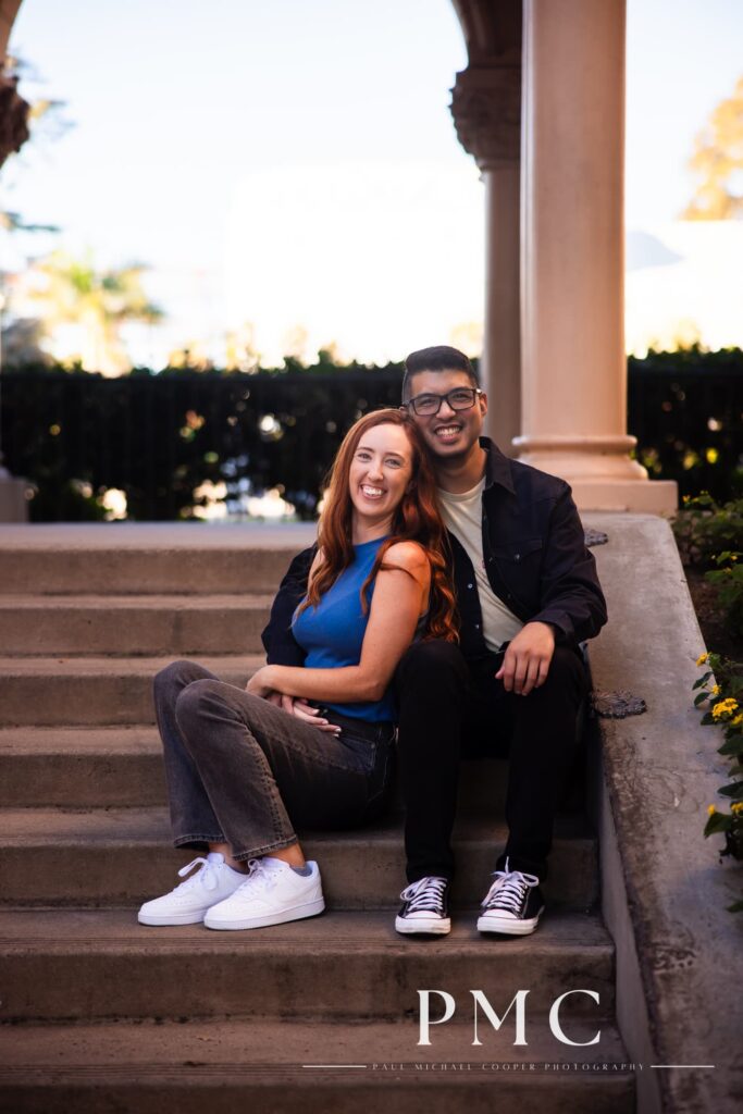 A couple smiles and sits on steps during their Balboa Park engagement session.