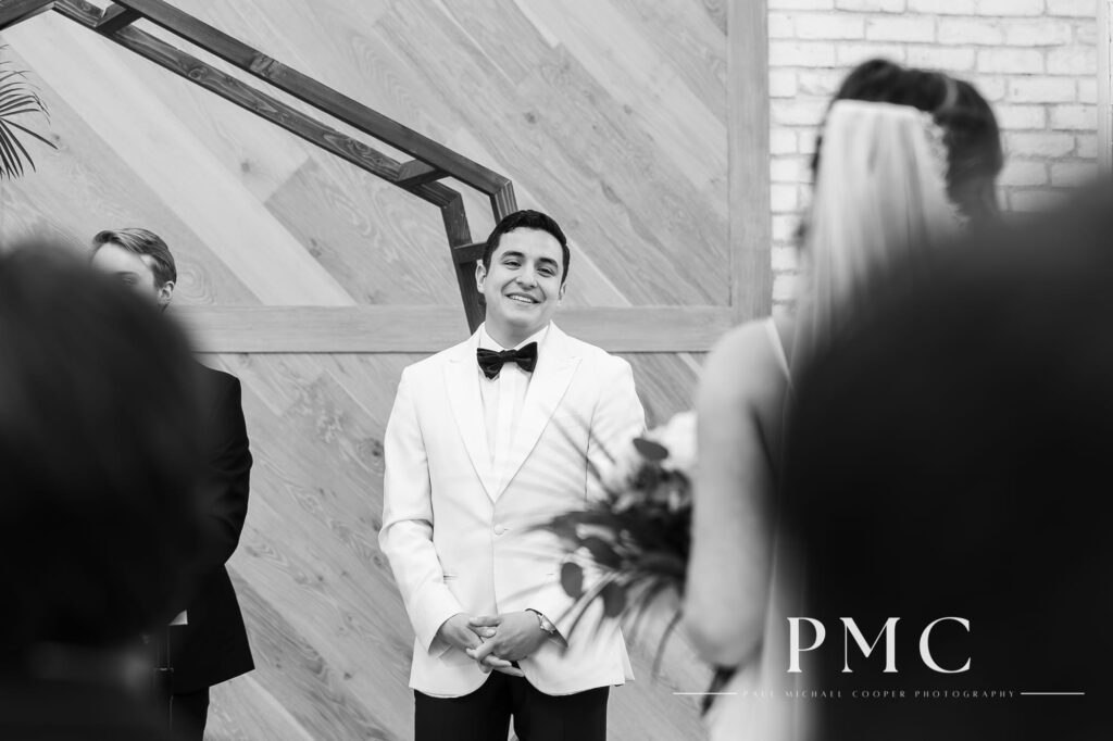 A black and white wedding photo of a groom smiling wide at his bride as she approaches him down the aisle at their chapel wedding ceremony.