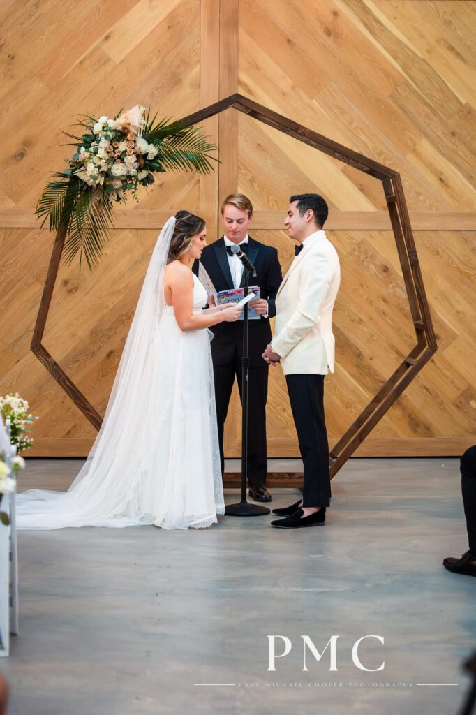 A bride with sweeping veil and her groom read their vows by a wooden arch with spring florals at their chapel wedding ceremony.