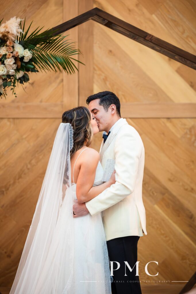 A bride and her groom seal their marriage with a kiss in front of their wooden arch with spring florals at their chapel wedding ceremony in Carlsbad, Southern California.