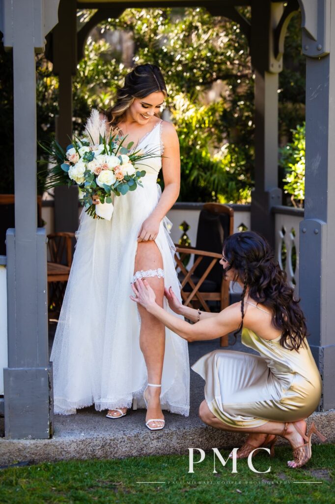 A bride holds her bouquet under a gazebo while her maid of honor adjusts her garter.