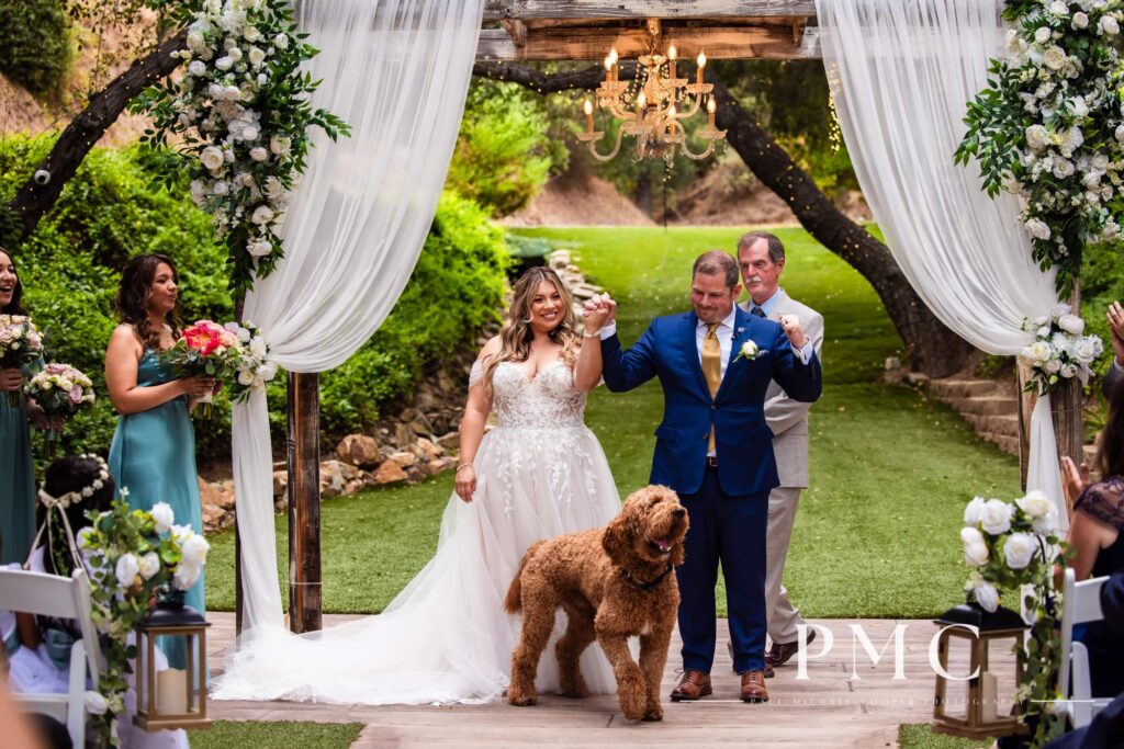 A bride and groom cheer beside their dog at their outdoor summer wedding ceremony.