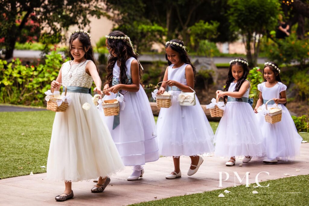 A line of 5 flower girls sprinkle petals on the ground at an outdoor summer wedding ceremony.