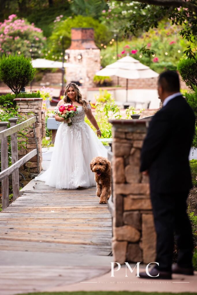 A bride with a vibrant floral bouquet enters her outdoor summer wedding ceremony with her beloved dog.