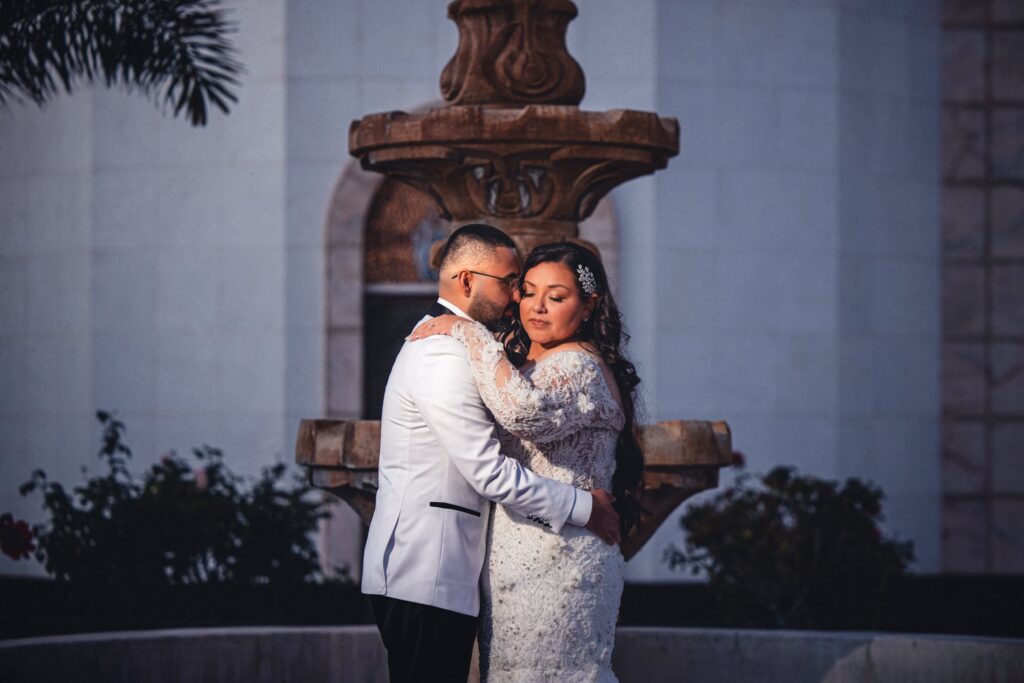 Portrait of a bride and groom embracing in front of a fountain, taken in a Classic wedding photography style.