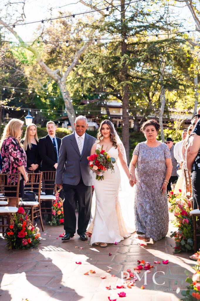 A bride is escorted by her parents down the aisle at her outdoor spring wedding.