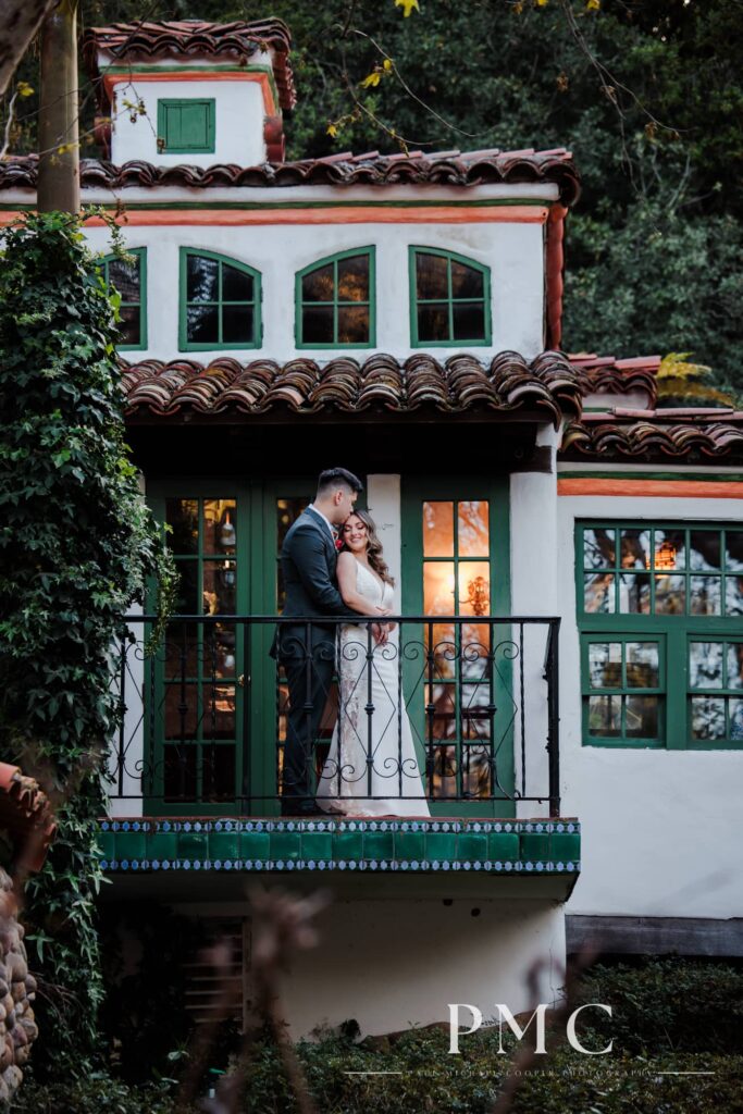 A bride and groom embrace each other on the balcony of a rancho-style building on their spring wedding day.