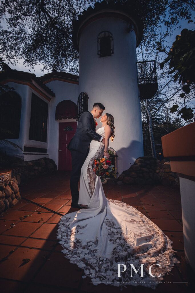 A bride and groom embrace in a dip with the bride's floral bouquet in front of a rancho-style building on their wedding day.