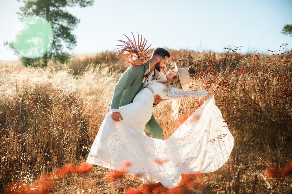 A trendy boho bride and groom with green suit and stylish hat doing a classic dip and dress train wedding photo in a field.