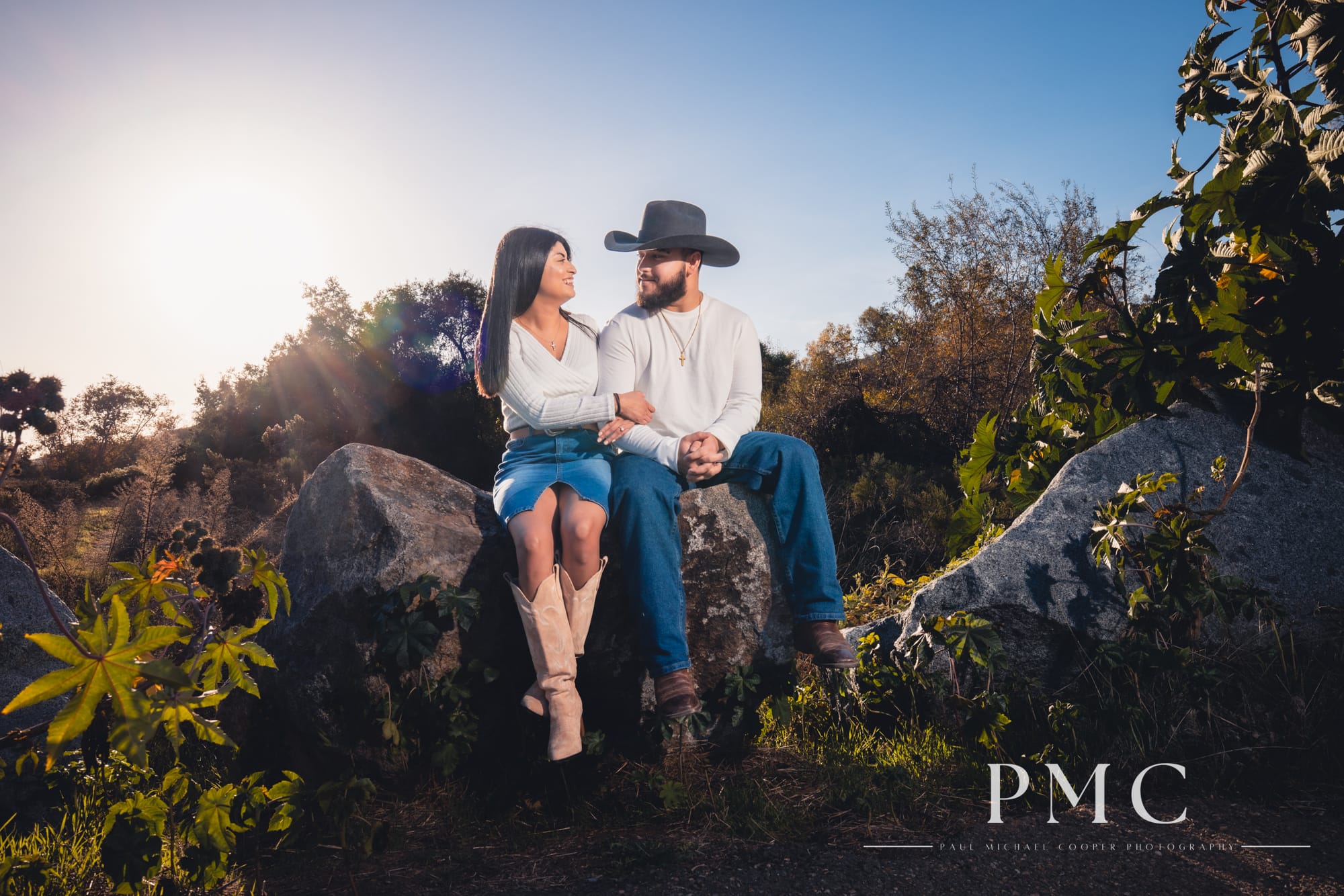 A couple smiles at each other while sitting on a rock during their fall engagement photos.