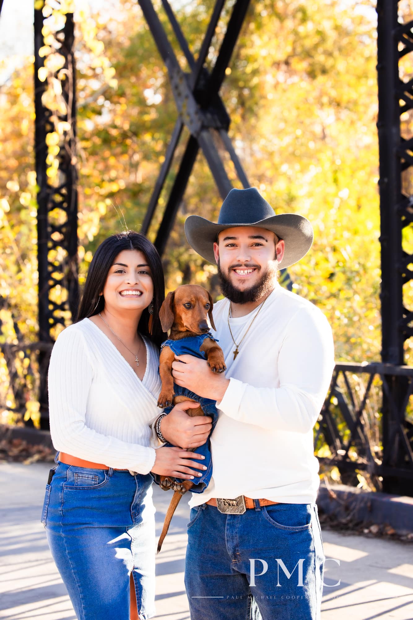 A couple smiles alongside their dog dressed in overalls in their fall engagement photos.