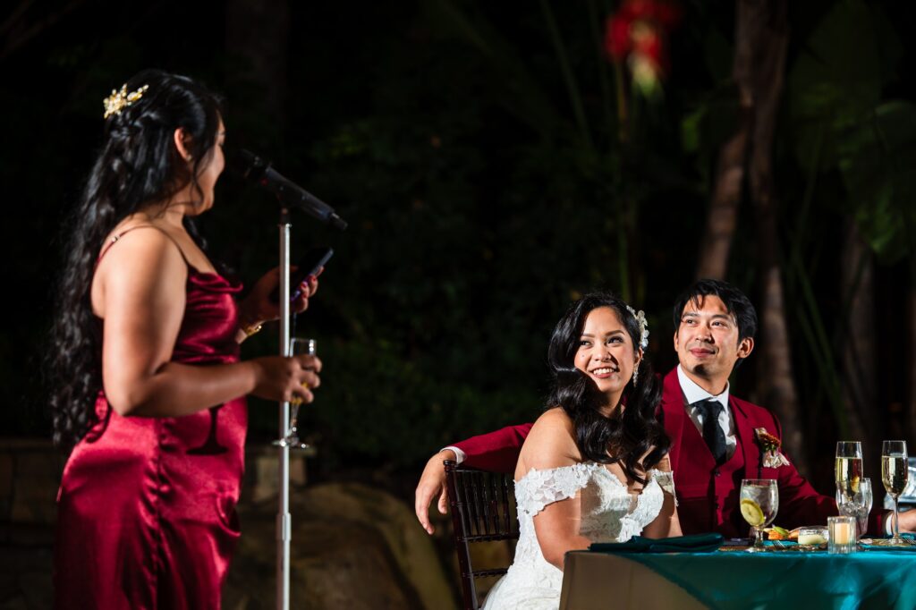 A bride and groom listening to a wedding toast given by the maid of honor.