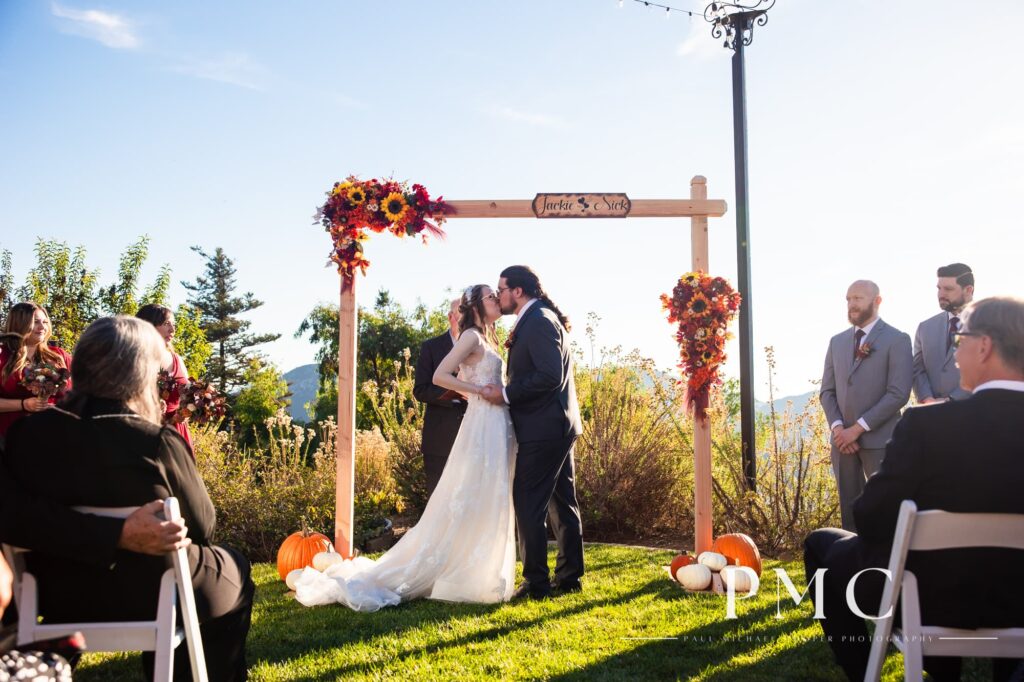 The bride and groom kiss beneath a custom arch with the couple's names and vibrant florals during their fall valley wedding in Escondido.