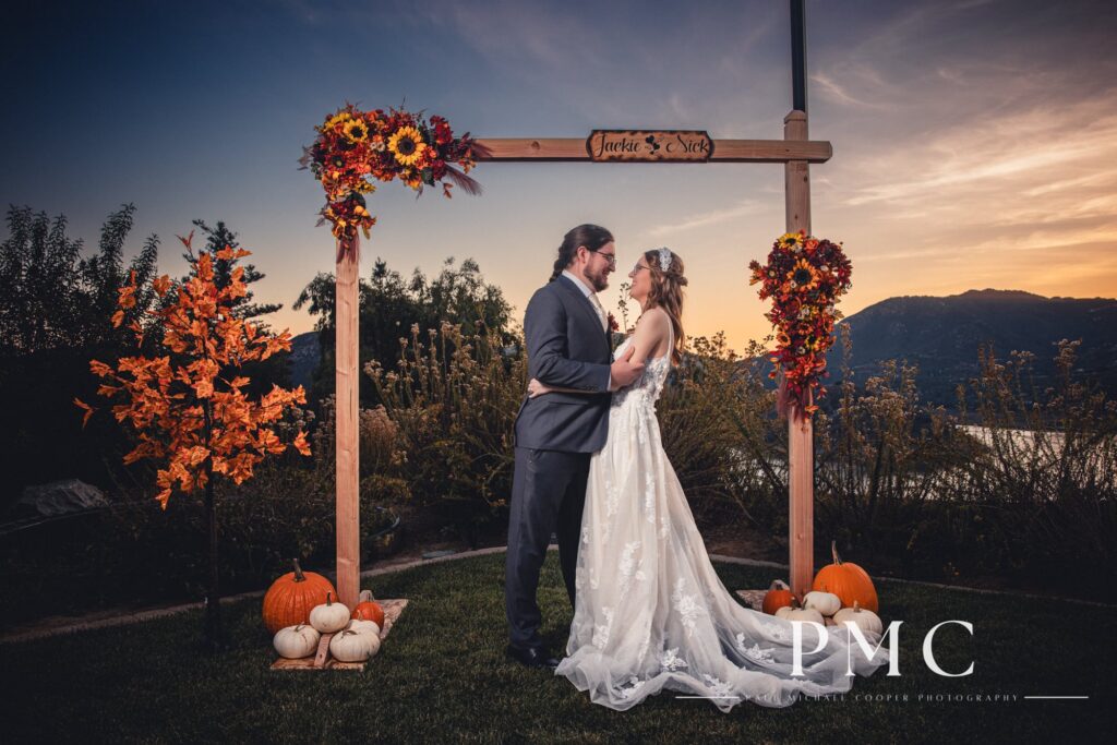 A bride and groom embrace each other under a custom arch with their names, surrounded by fall flowers and decor at sunset at their wedding overlooking the Escondido valley.