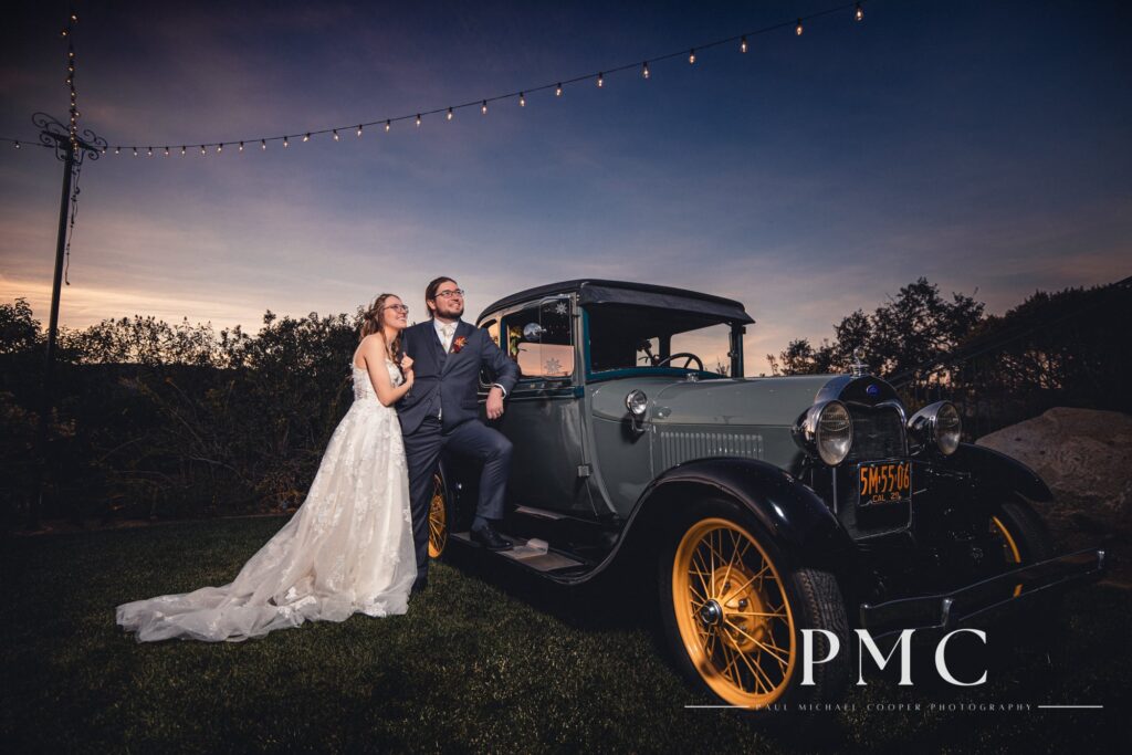 A bride and groom pose with a vintage car at sunset at their fall valley wedding on a private estate in Escondido.