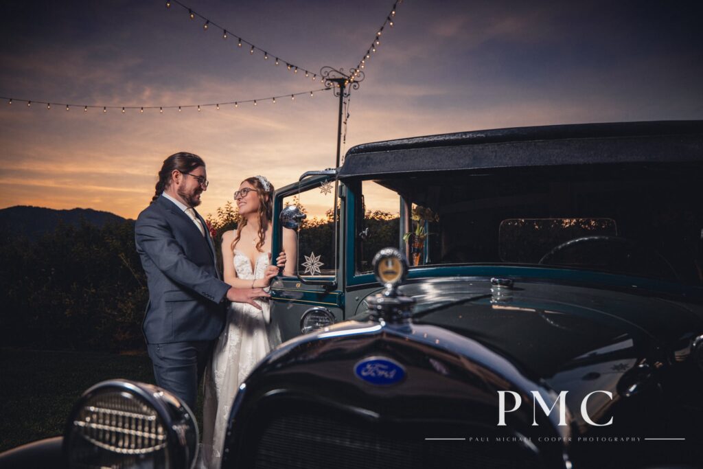 A bride and groom pose with a vintage car at sunset at their fall valley wedding on a private estate in Escondido.