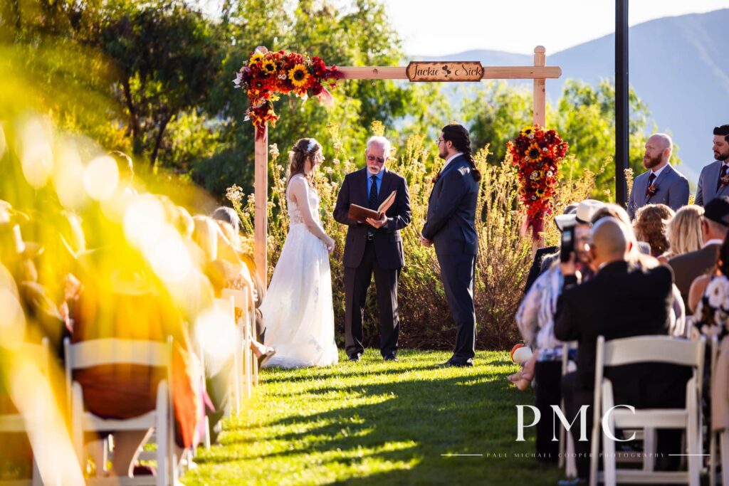 A photo of the ceremony, with a custom arch with the couple's names and vibrant florals, at a fall valley wedding in Escondido.
