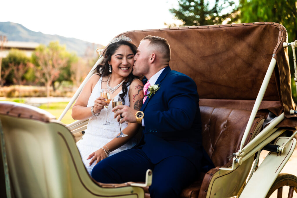 A newlywed bride and groom toast each other with champagne flutes and share a kiss in a horse-drawn carriage after their outdoor wedding ceremony at Galway Downs by Wedgewood Weddings in Temecula.