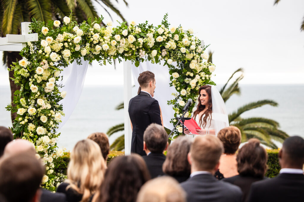 A bride reads her vows to her groom under an arch of white roses at their outdoor wedding ceremony on the lawn of the Bel-Air Bay Club in Los Angeles.