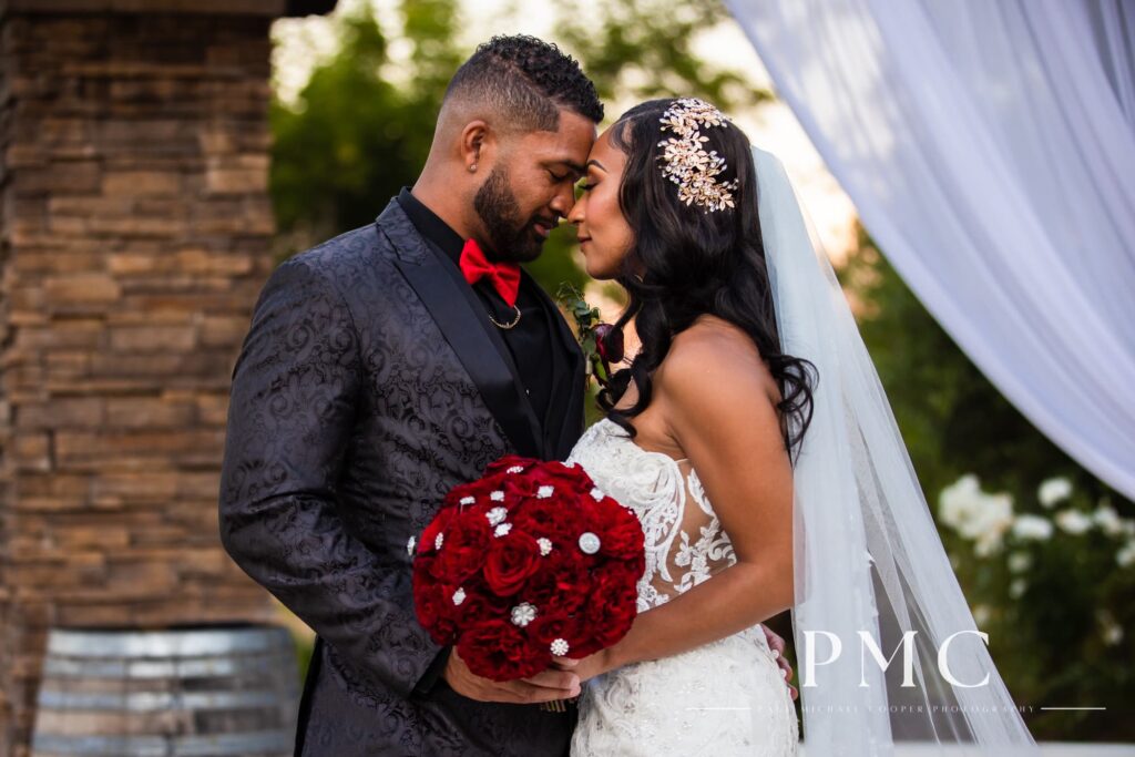 A bride and groom stand together in an embrace with a vibrant red floral bouquet on their wedding day at Fallbrook Estate in Southern California.