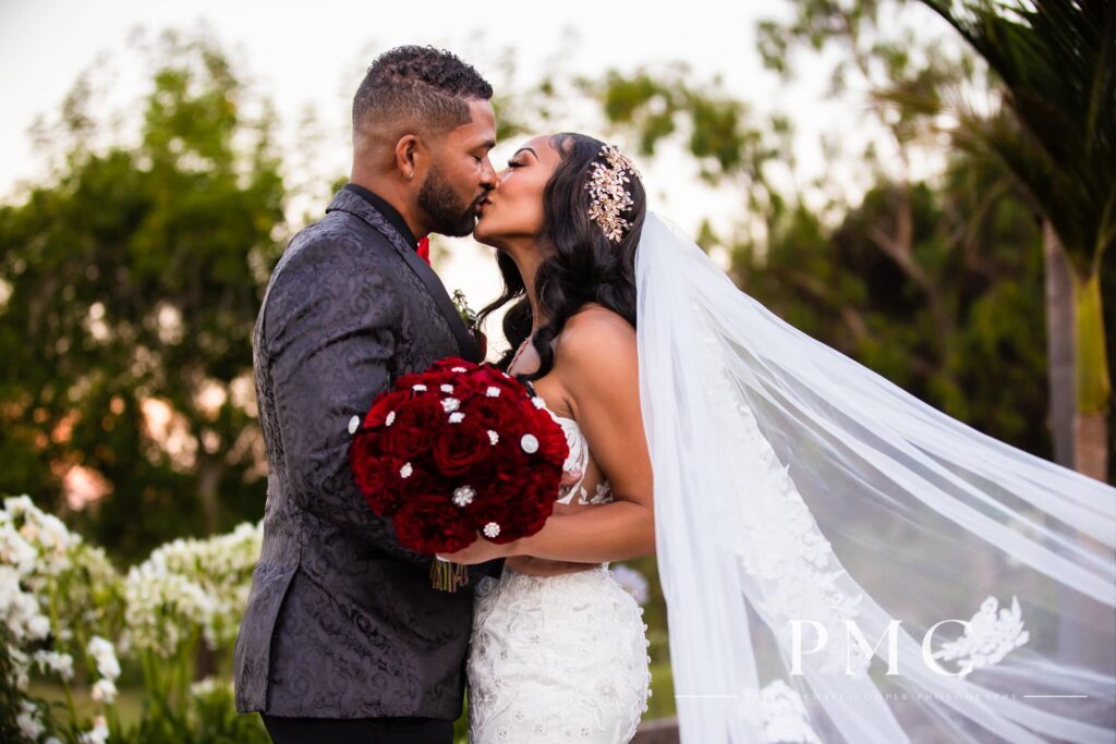 A bride and groom share a kiss in an embrace with a vibrant red floral bouquet and sweeping bridal veil on their wedding day at Fallbrook Estate in Southern California.