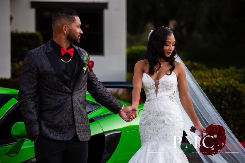 A bride and groom walk hand in hand with a vibrant red floral bouquet and sweeping bridal veil in front of a bright green McLaren wedding car.