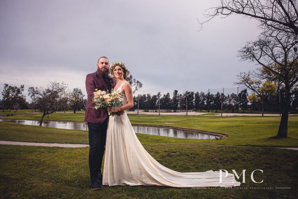 A groom in a maroon suit jacket and his bride, who is wearing her grandmother's vintage wedding dress and a boho floral crown, smile for the camera in front of a lake at the David L. Baker Golf Course.