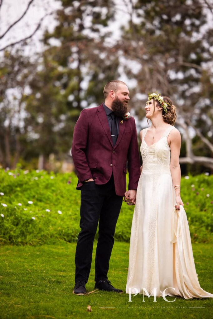 A groom in a maroon suit jacket and bride in a vintage wedding dress and floral crown hold hands and smile at each other in nature.