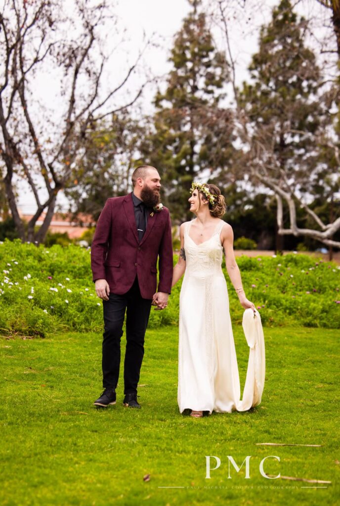 A groom in a maroon suit jacket and bride in a vintage wedding dress and floral crown hold hands and smile at each other in nature in Orange County.