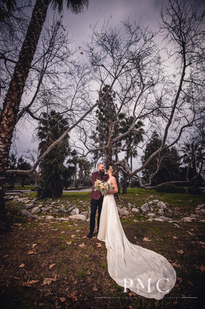 A groom in a maroon suit jacket and bride in a vintage wedding dress and floral crown hold a boho-inspired floral bouquet and look at each other in nature in Orange County.