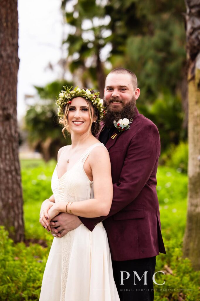 A groom in a maroon suit jacket and bride in a vintage wedding dress and floral crown hold each other in nature in Orange County.