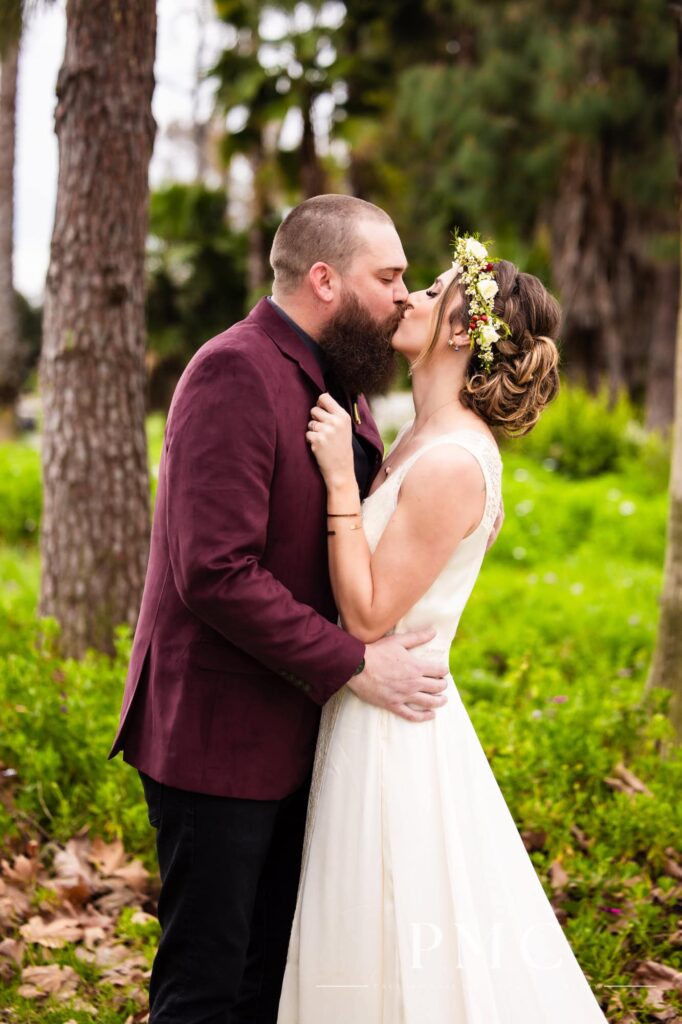A groom in a maroon suit jacket and bride in a vintage wedding dress and floral crown hold each other and kiss in nature in Orange County.