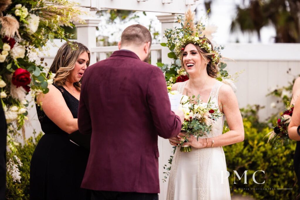 A bride in a vintage wedding dress and boho-inspired floral crown smiles as her groom reads his vows during their Orange County wedding ceremony.
