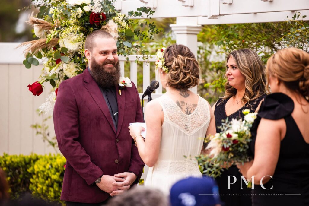 A groom in a vintage maroon suit jacket smiles at his bride in a vintage wedding dress reading her vows during their Orange County wedding ceremony.