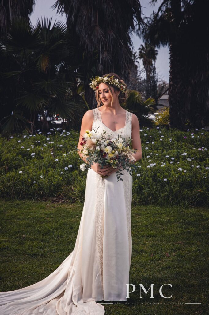 A bride wears her grandmother's vintage wedding dress and boho floral crown while holding a flower bouquet among greenery and trees.