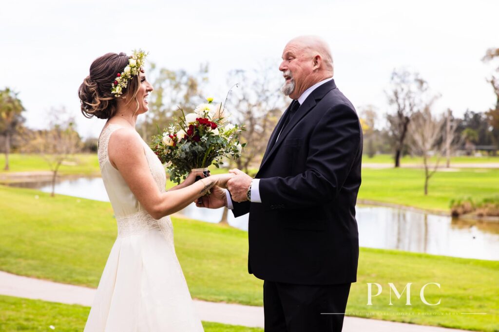 A bride in her grandmother's vintage wedding dress and boho floral crown shares a happy First Look moment with the father of the bride, overlooking a lake.
