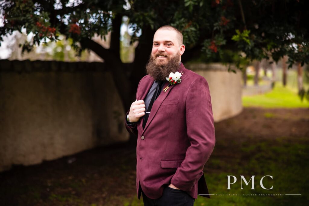 A groom smiles at the camera, wearing a vintage maroon jacket.