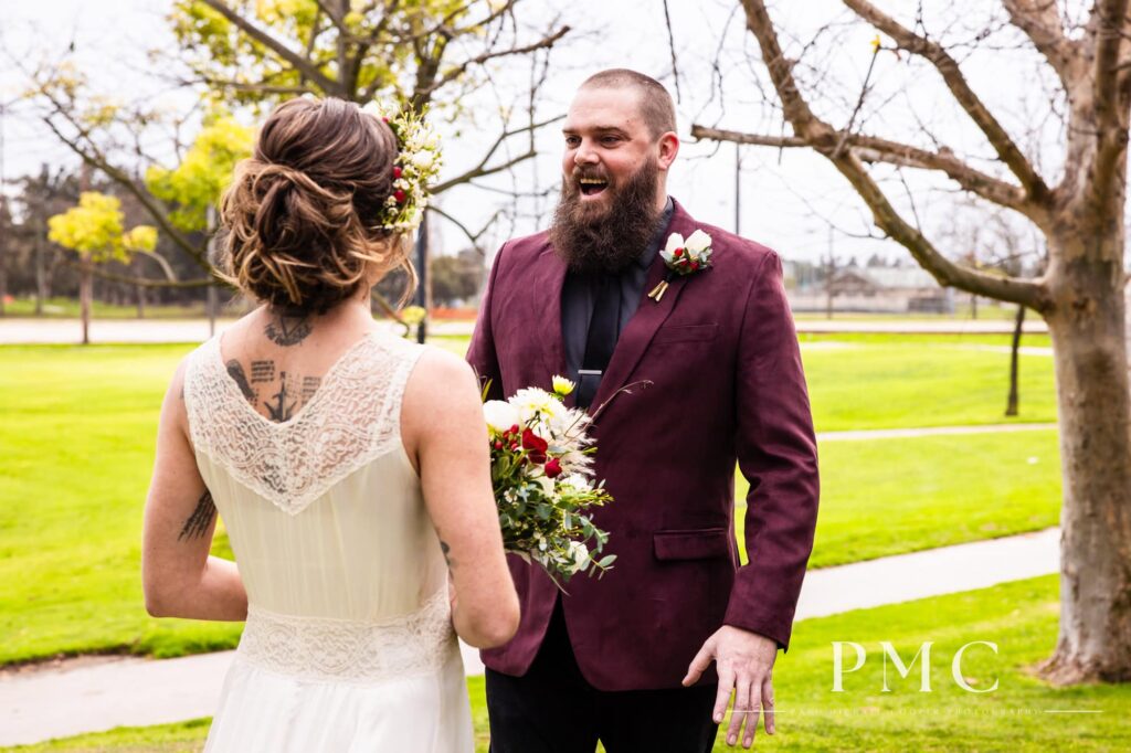 A groom in a maroon suit jacket smiles excitedly during his First Look with his bride, who is wearing her grandmother's vintage wedding dress and a boho floral crown.