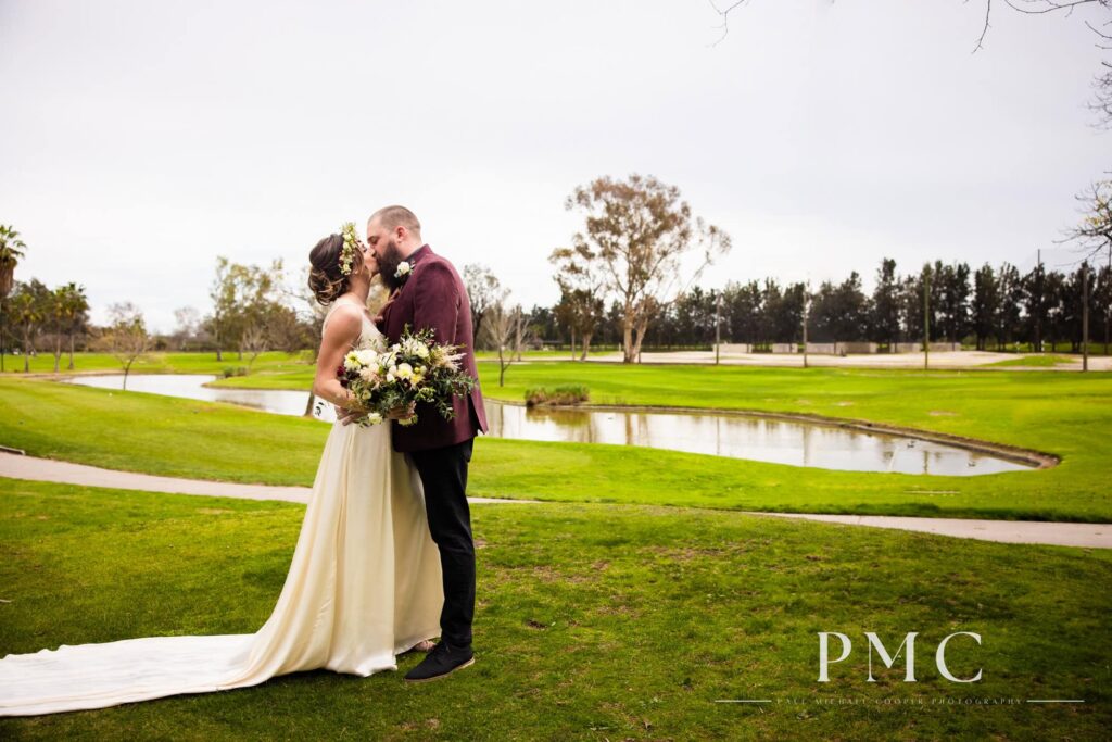 A groom in a maroon suit jacket shares a kiss with his bride, who is wearing her grandmother's vintage wedding dress and a boho floral crown, overlooking a lake.