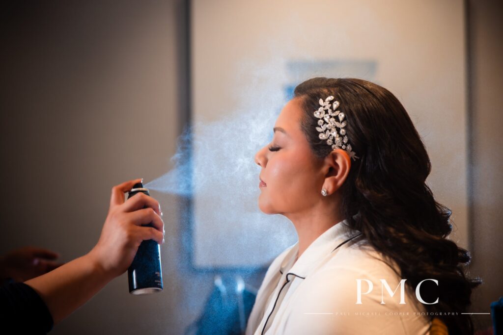 A bride in a white robe with a jeweled hairpiece gets her makeup setting spray on her wedding day.